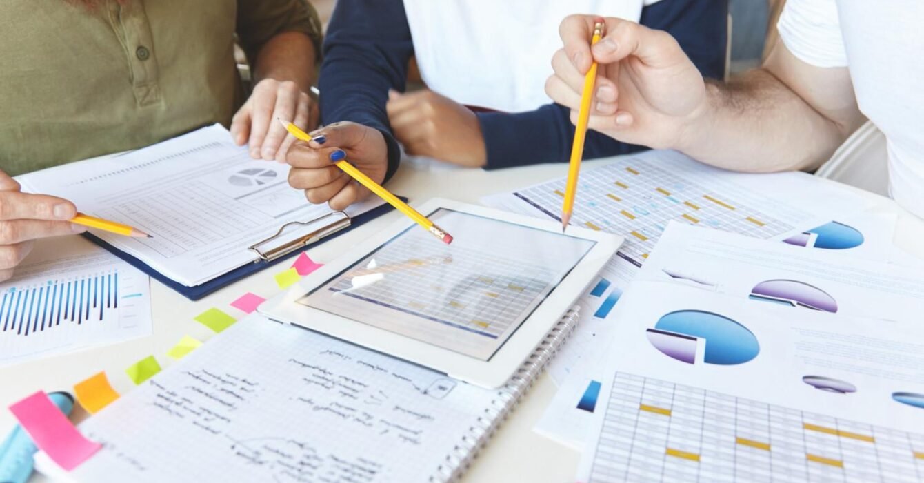 Syria Feasibility Study: A diverse group of people analyzing charts and data on a tablet and papers, with pencils and sticky notes scattered on a table.